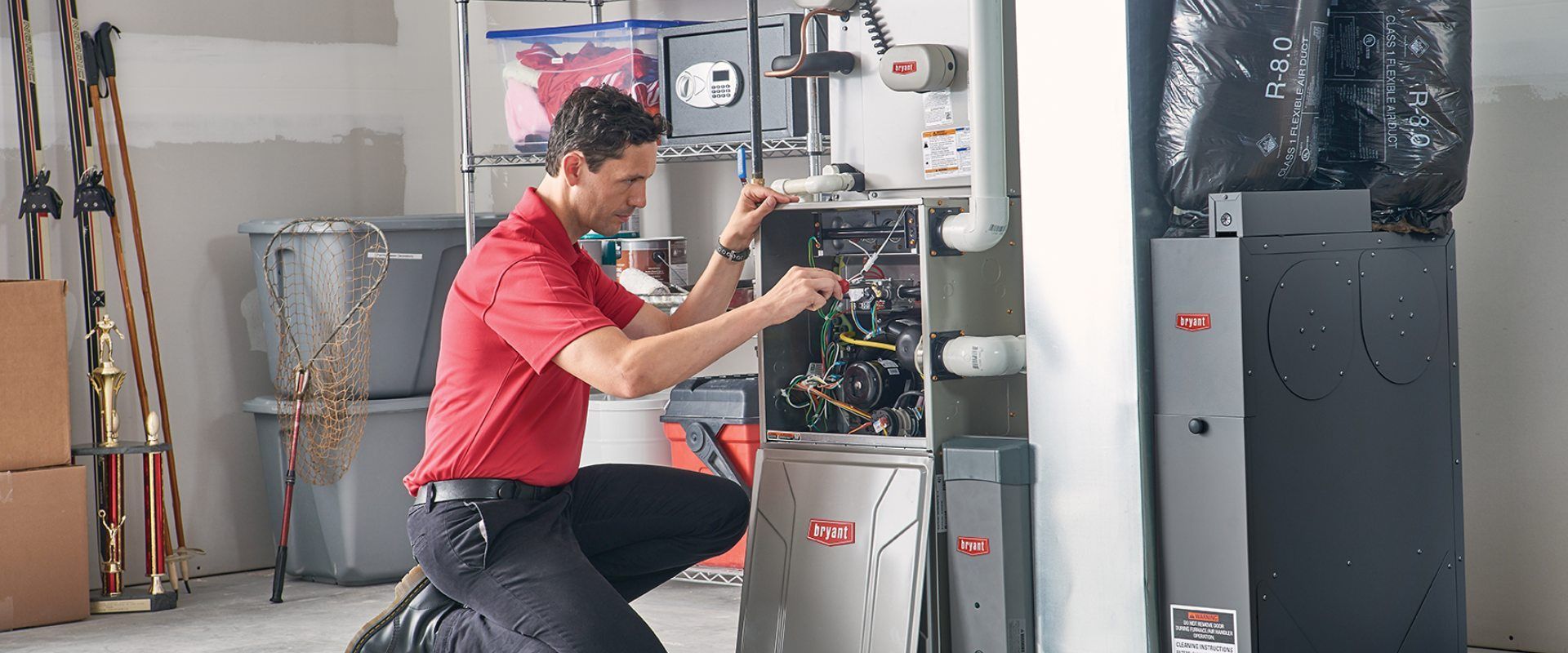 A man is kneeling down in front of a furnace in a garage.