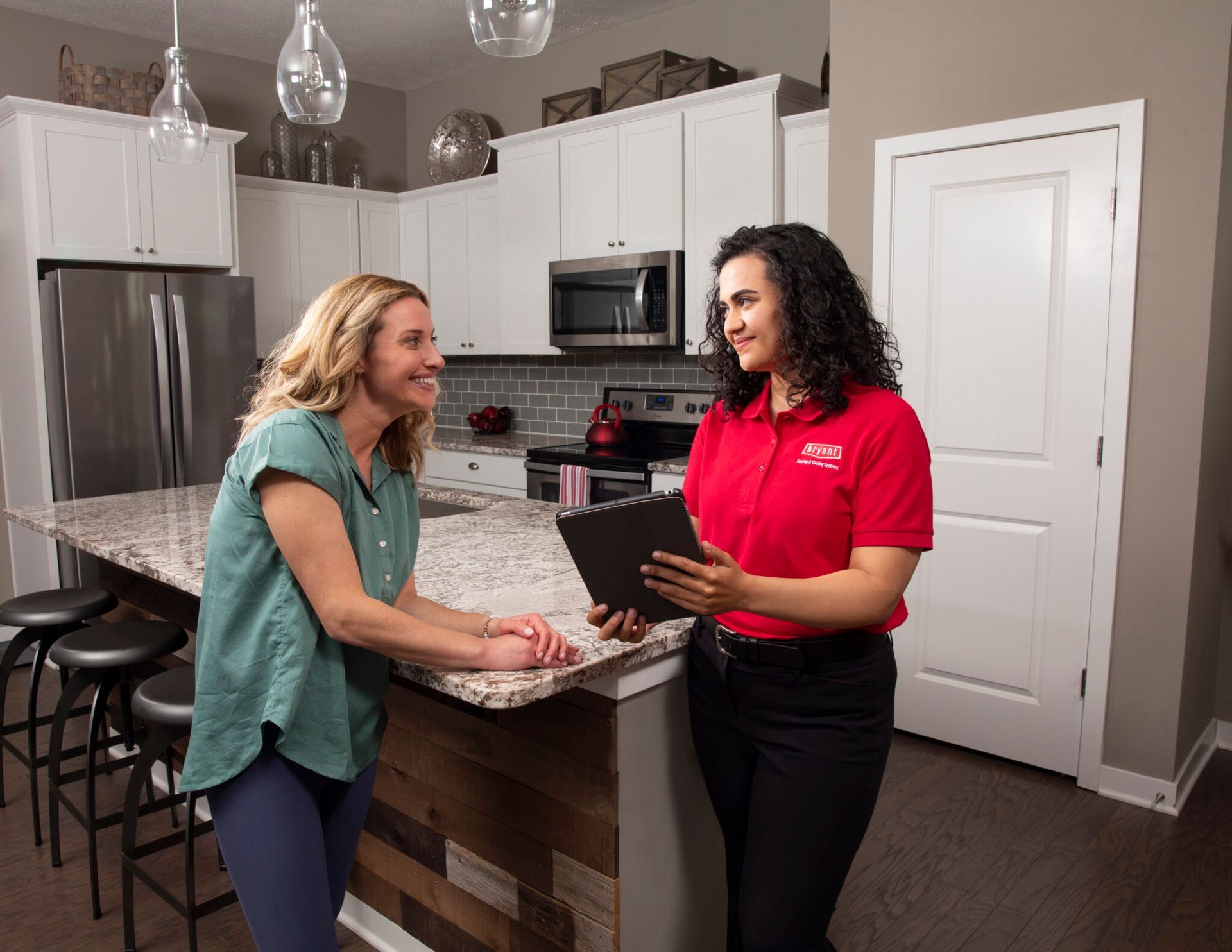 A woman in a red shirt is talking to another woman in a kitchen.