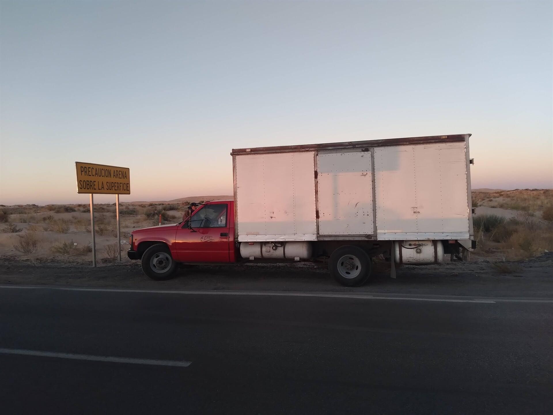 Un camión rojo con una caja blanca en la parte trasera está estacionado al costado de la carretera.