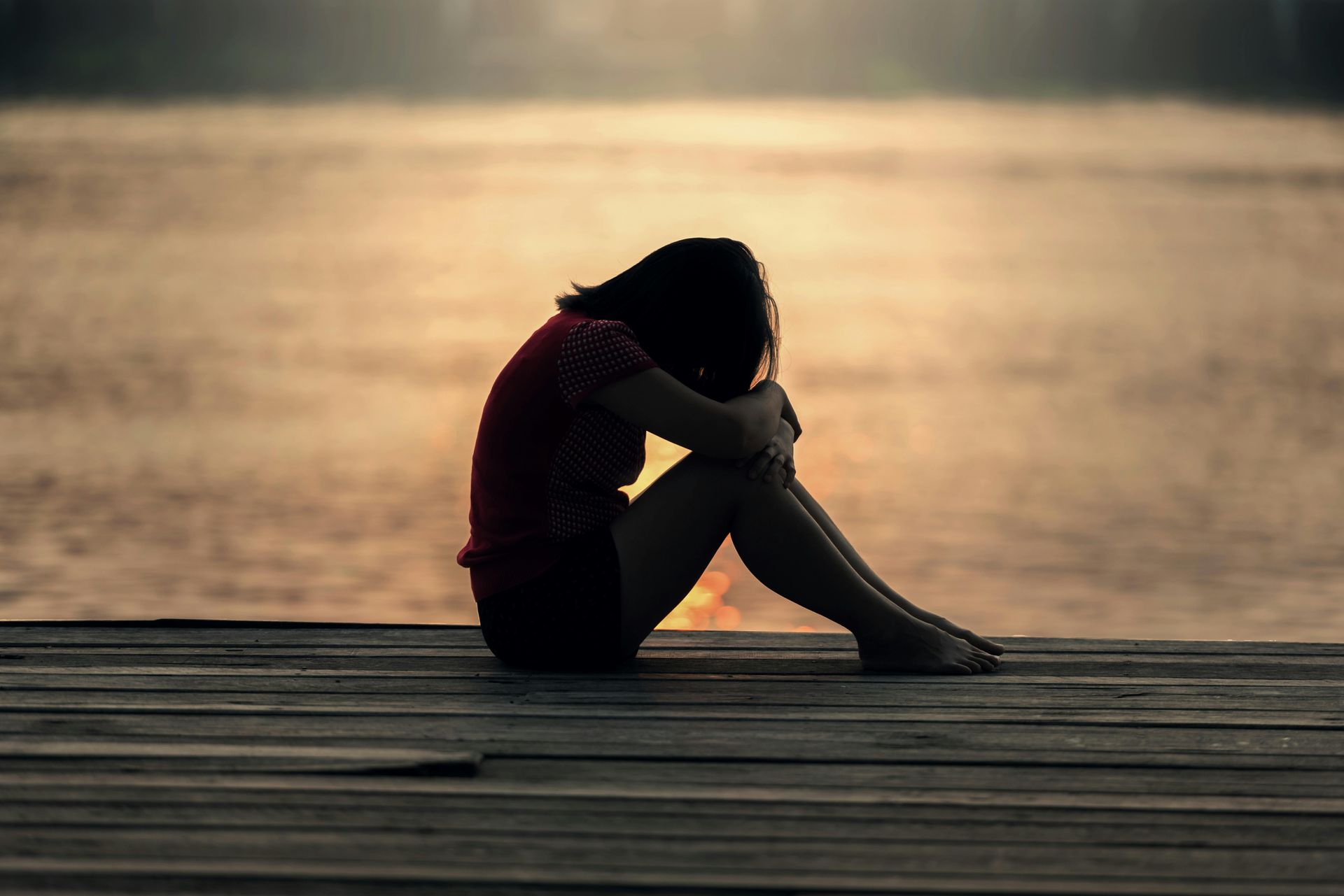 Woman sitting alone on a dock, head in arms, overlooking water during sunset.