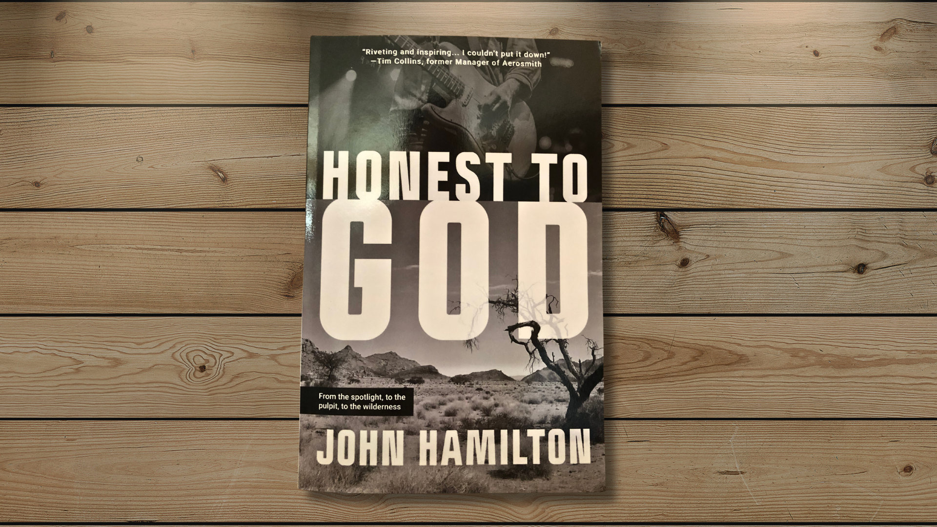 The cover of a paperback book against a background of wood planks. The cover features the title, Honest to God, in large white, all-caps letters across the center of the book. The author's name, John Hamilton, is in a slightly smaller font, all caps, across the bottom of the cover. The cover's background is grayscale and shows a man playing guitar on stage in the top third and a desolate scene of a dead tree in front of scrubland leading to barren mountains in the bottom third.