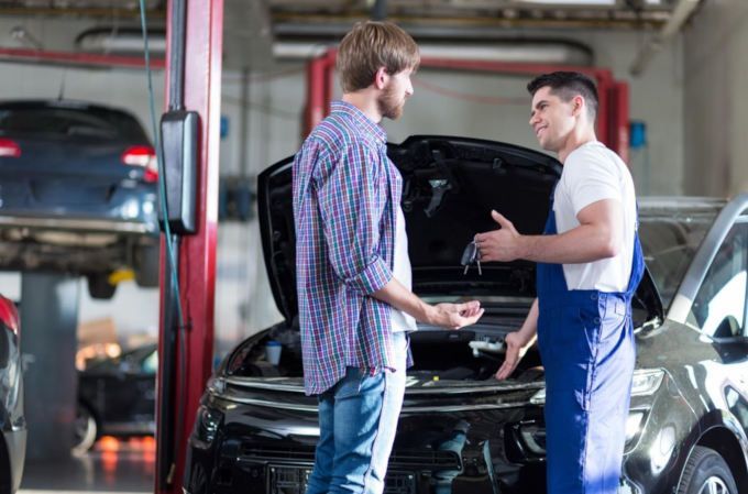 Two mechanics talking beside a car in an auto repair shop, with the hood open.