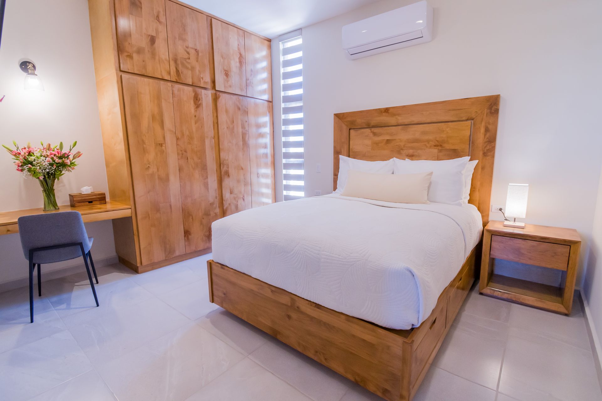 Dormitorio luminoso y minimalista con cama de madera, armario, mesita de noche, silla y paredes blancas.