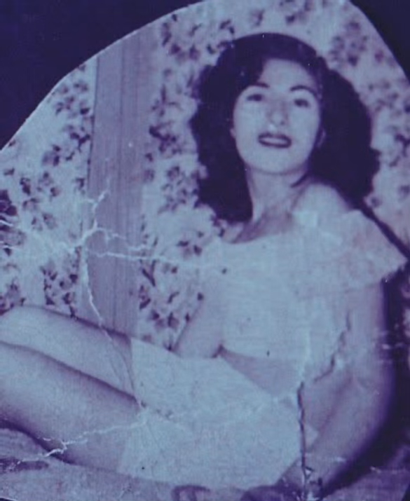 Woman in a white dress, seated against a floral background; smiling with dark hair and dark lipstick.