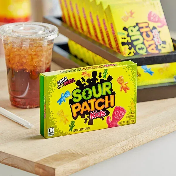 Box of Sour Patch Kids candy on a wooden board, a drink, and other boxes in background.