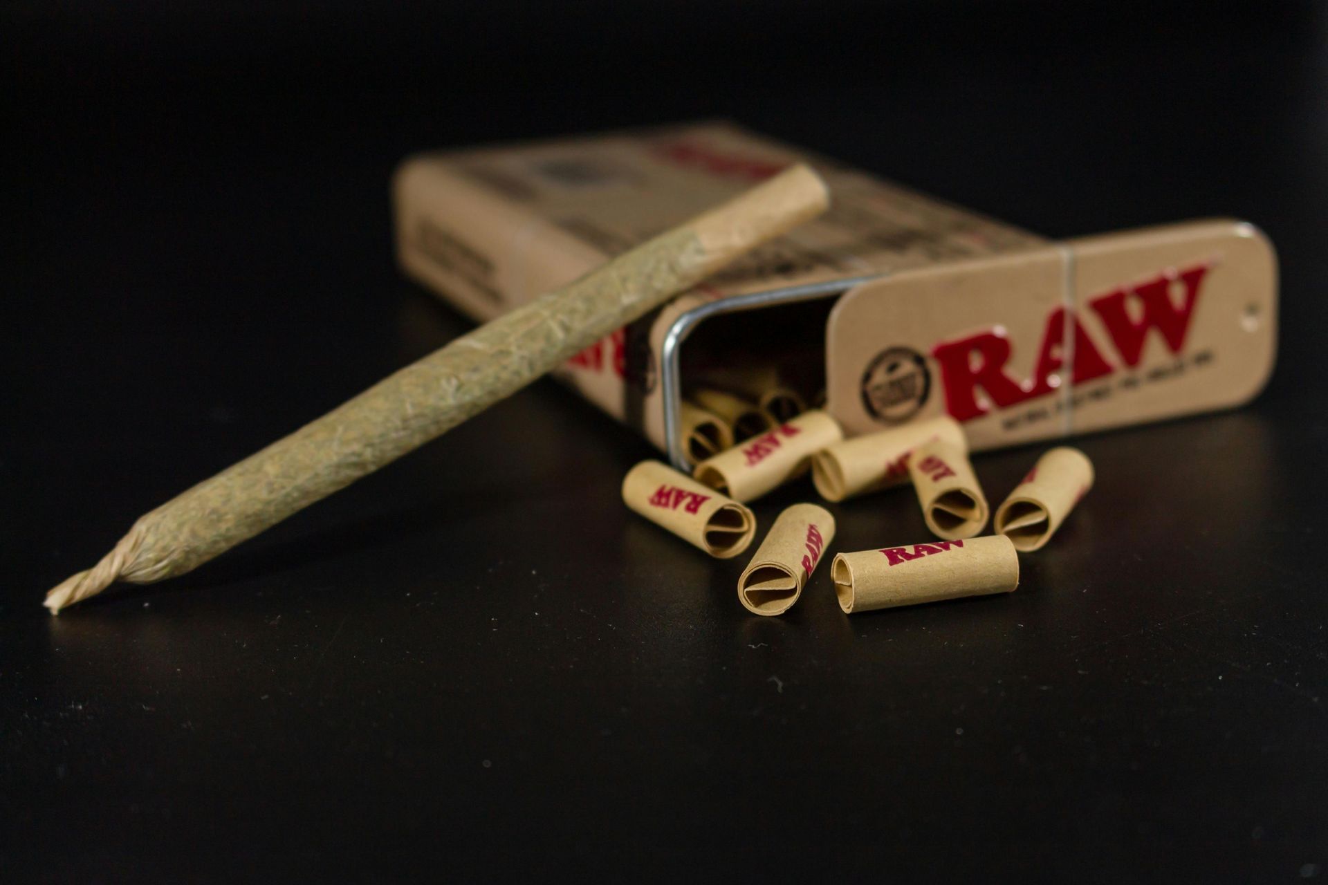 A rolled cannabis joint next to an open RAW rolling paper tin and filter tips on a dark surface.