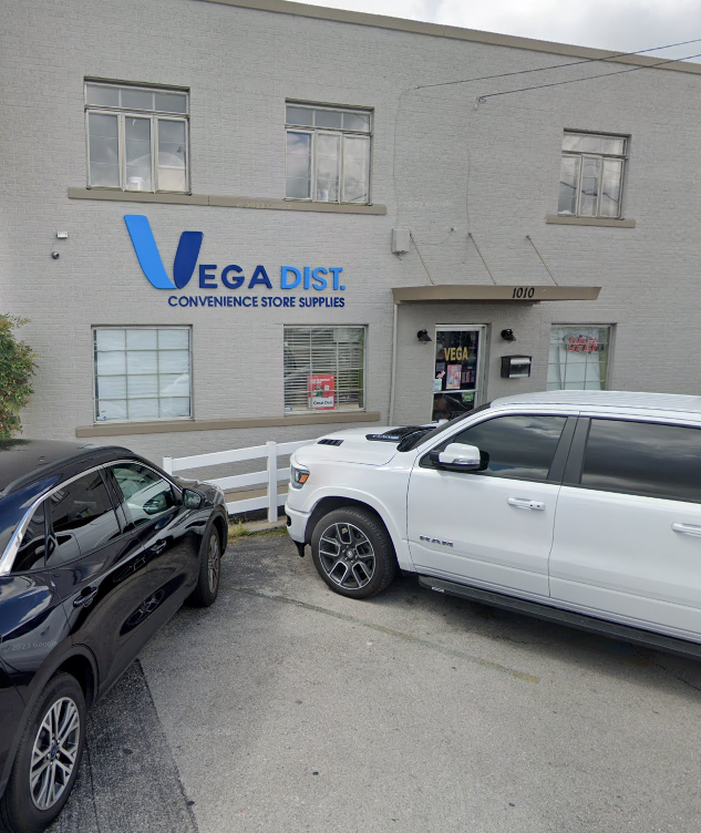 Exterior of Vega Dist Convenience Store Supply building with a white SUV and black car parked outside.