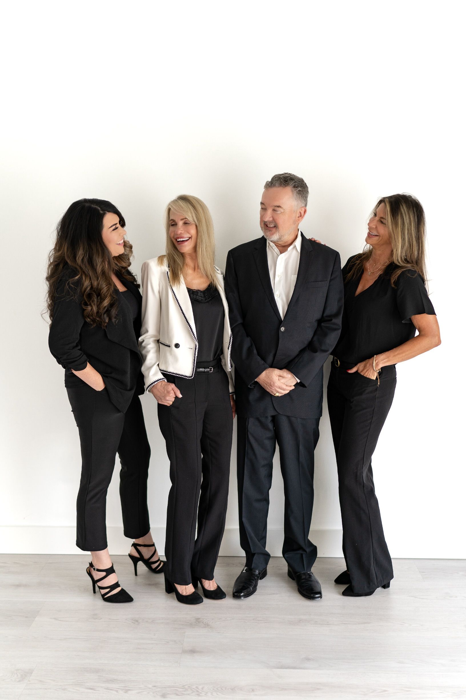 Four people stand against a white wall. Two women on either side of a man, all smiling. One woman in black with heels, one in a white blazer and black pants. The man in a dark suit.