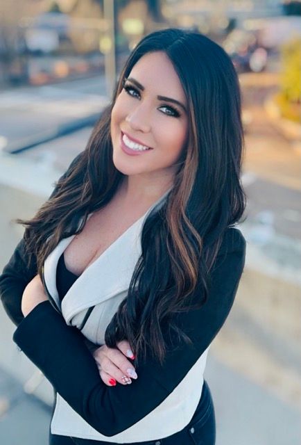 A woman in a black and white jacket is standing with her arms crossed and smiling.