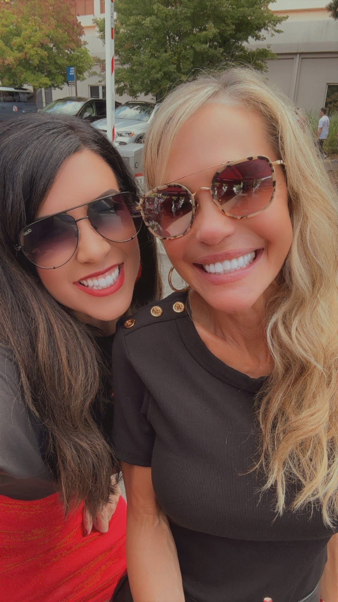 Two smiling women wearing sunglasses. One with dark hair, the other blonde, near a street.