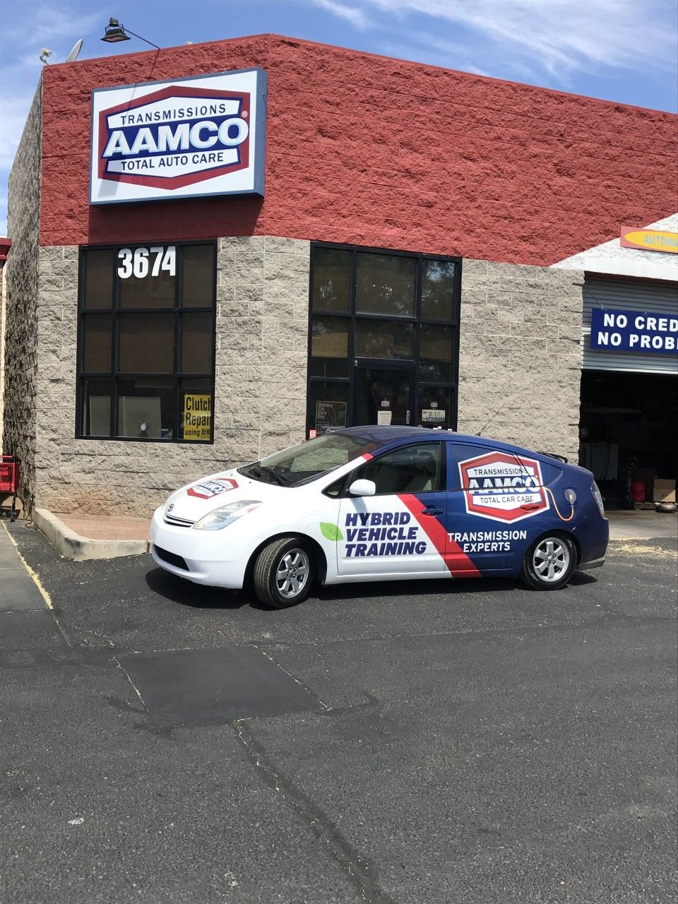 AAMCO Tucson, Az (Golf Links Rd) transmission shop with a hybrid vehicle training car parked outside. Building has a red and gray brick exterior.