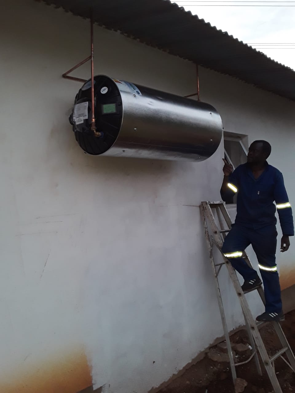 worker standing on a ladder and inspecting a  wall mounted electric geyser
