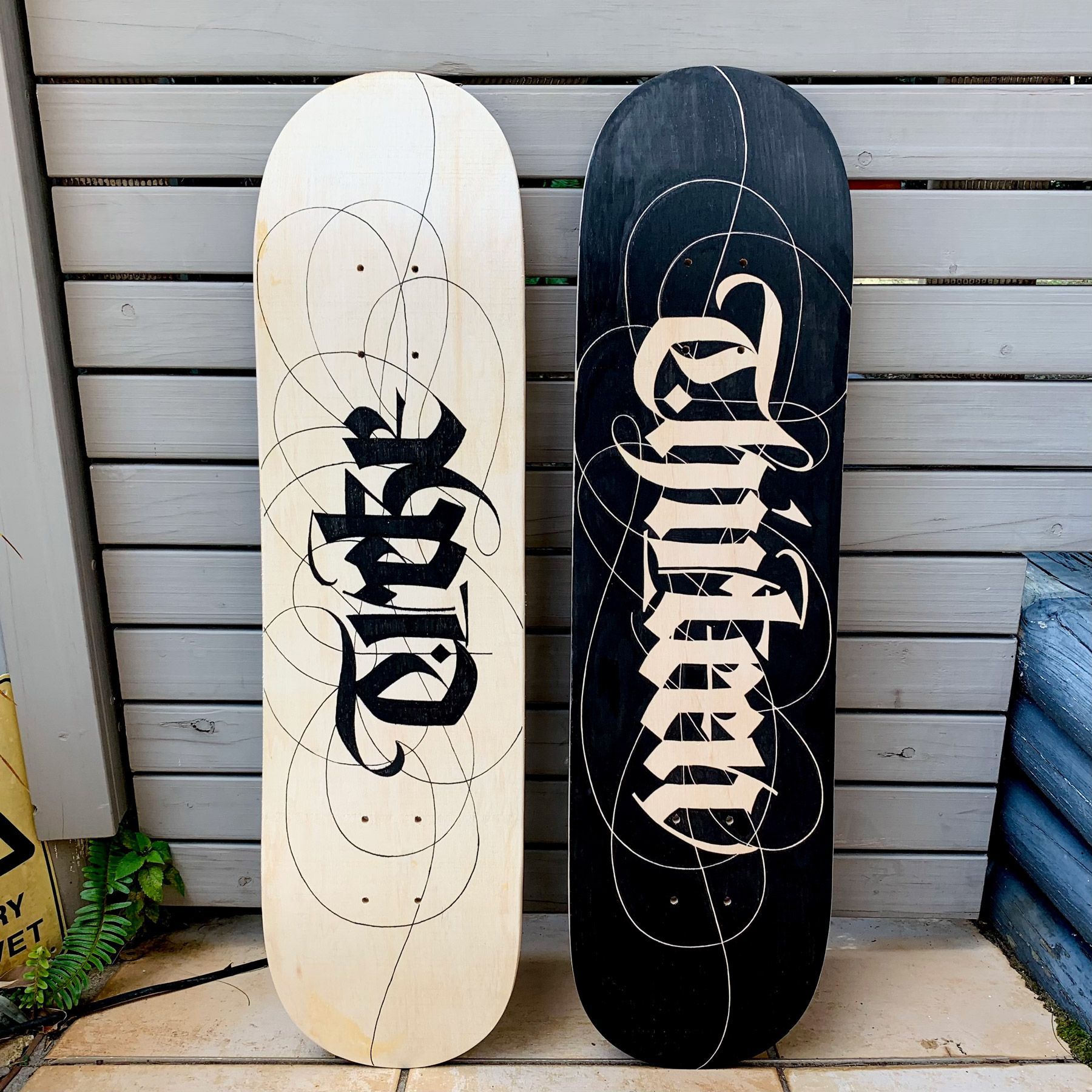 Two skateboard decks lean against a grey wood wall; one is light wood with black script, the other black with white script.