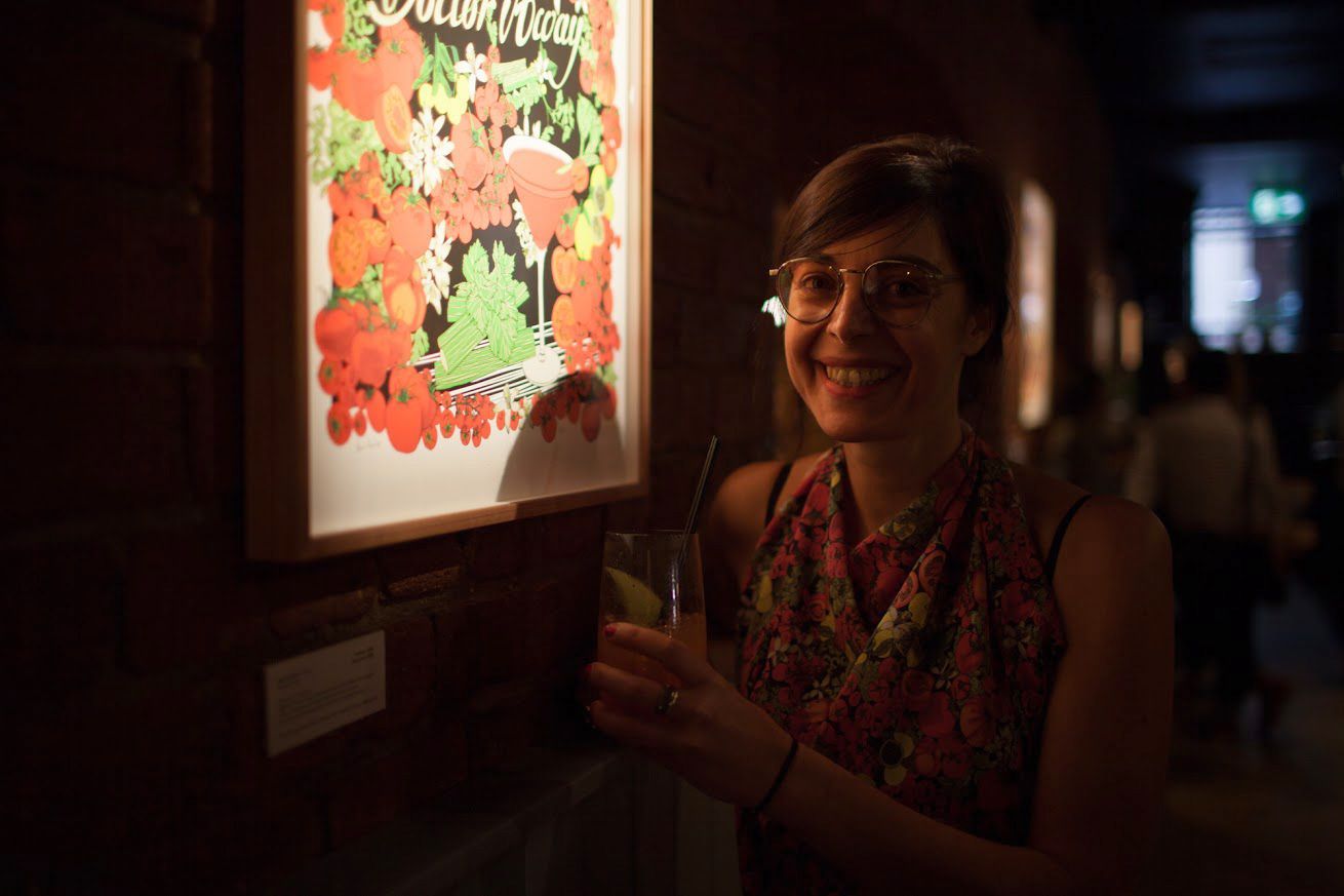 Maria Montes smiling beside a brightly lit floral artwork in a dim gallery hallway