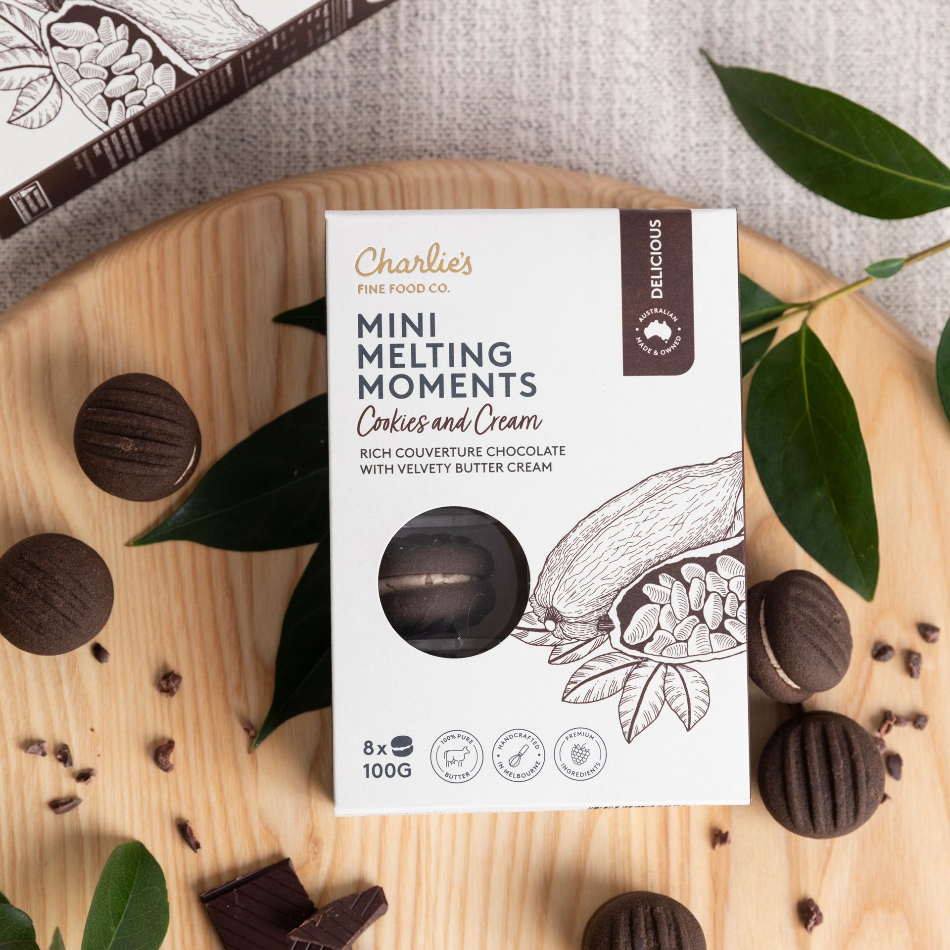 Box of Charlie’s Mini Melting Moments biscuits on a wooden board with chocolate cookies and green leaves
