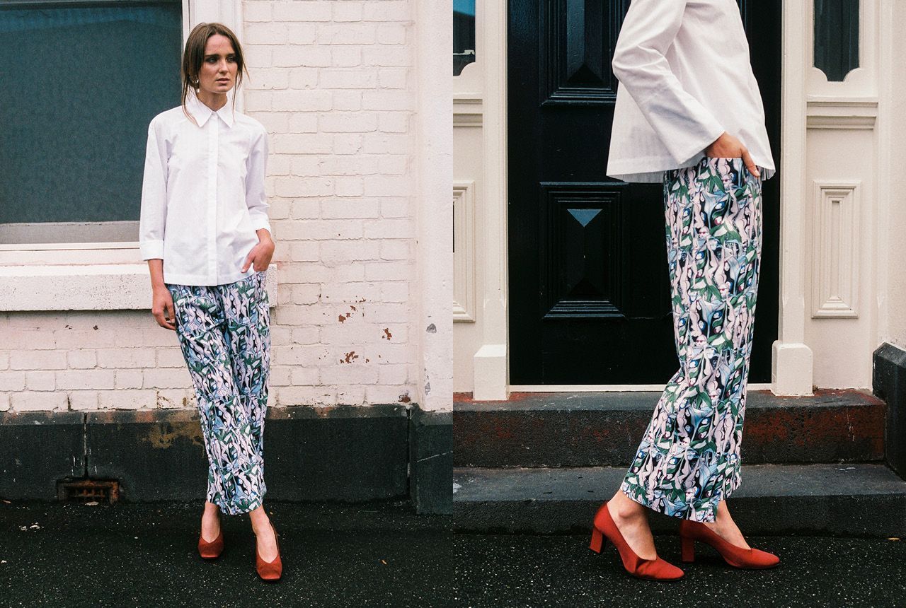 Person in a white shirt and patterned pants stands on a sidewalk beside a building.