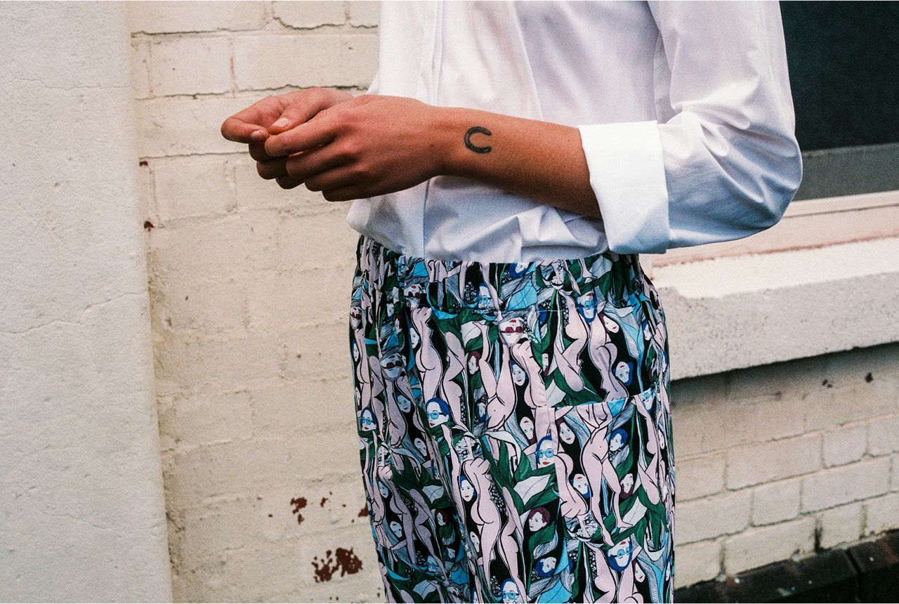 Person in white shirt and patterned skirt standing by a brick wall, holding a small object.