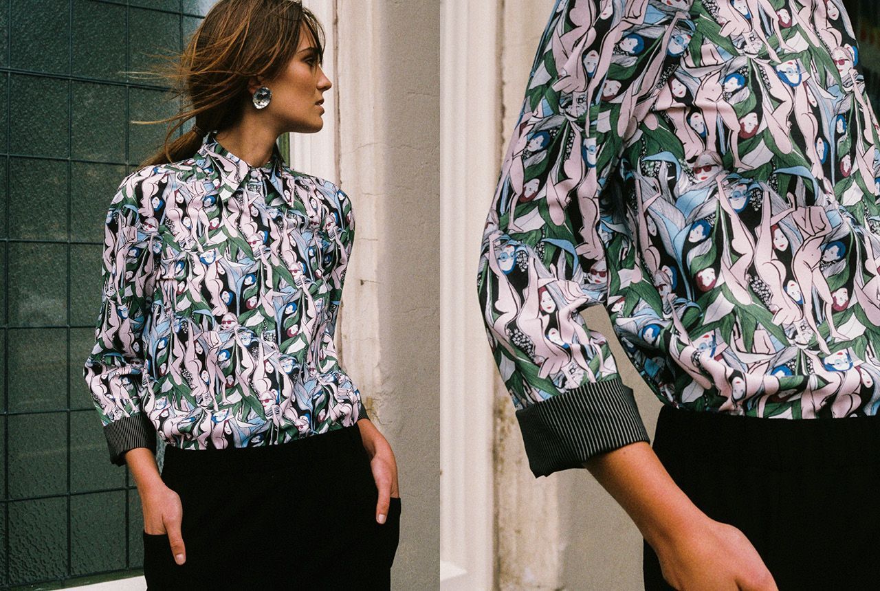 Person wearing a patterned blouse and black pants, shown standing in profile and close-up near a wall.