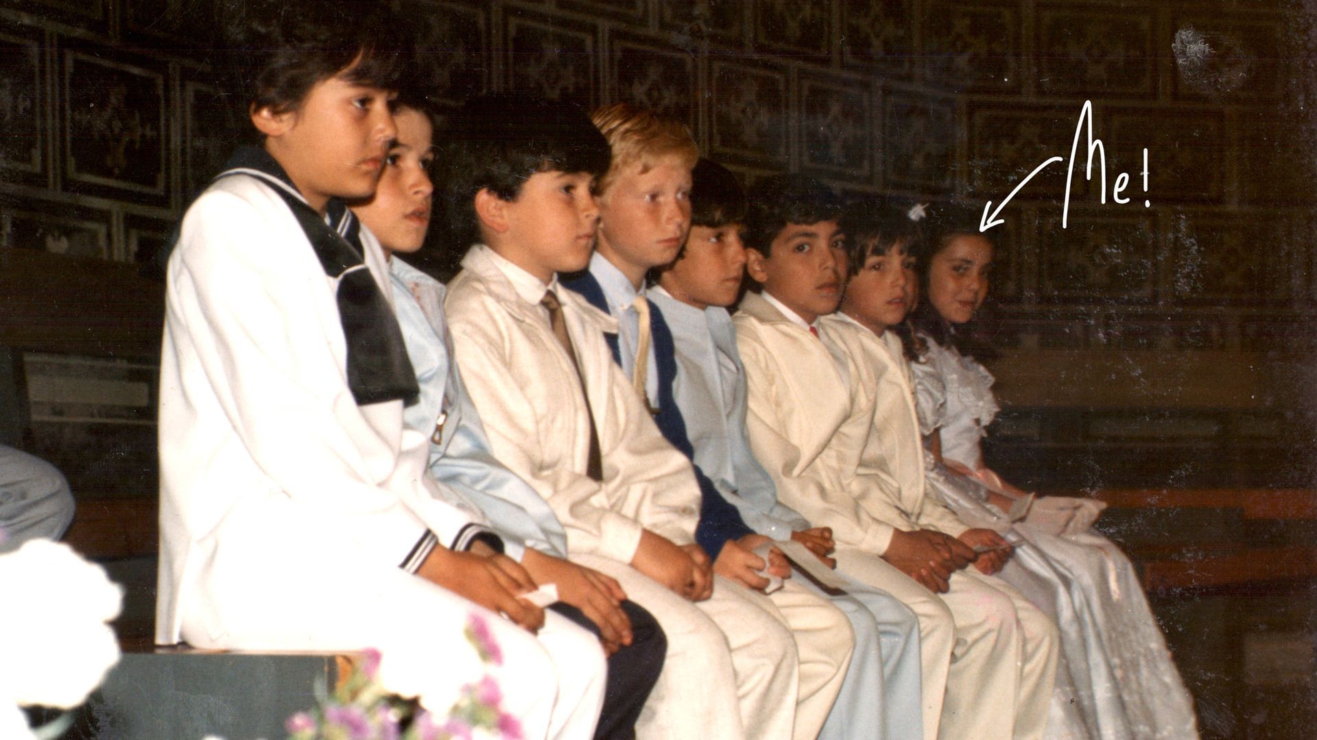 A row of children in formal white attire sit side-by-side in a dimly lit room, with an arrow pointing to one person.