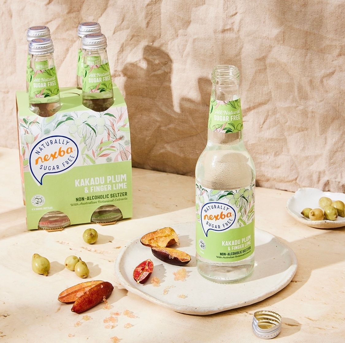 Glass bottle and two-pack of green-labeled kiwi pear drink on a white plate with sliced fruit on a beige backdrop