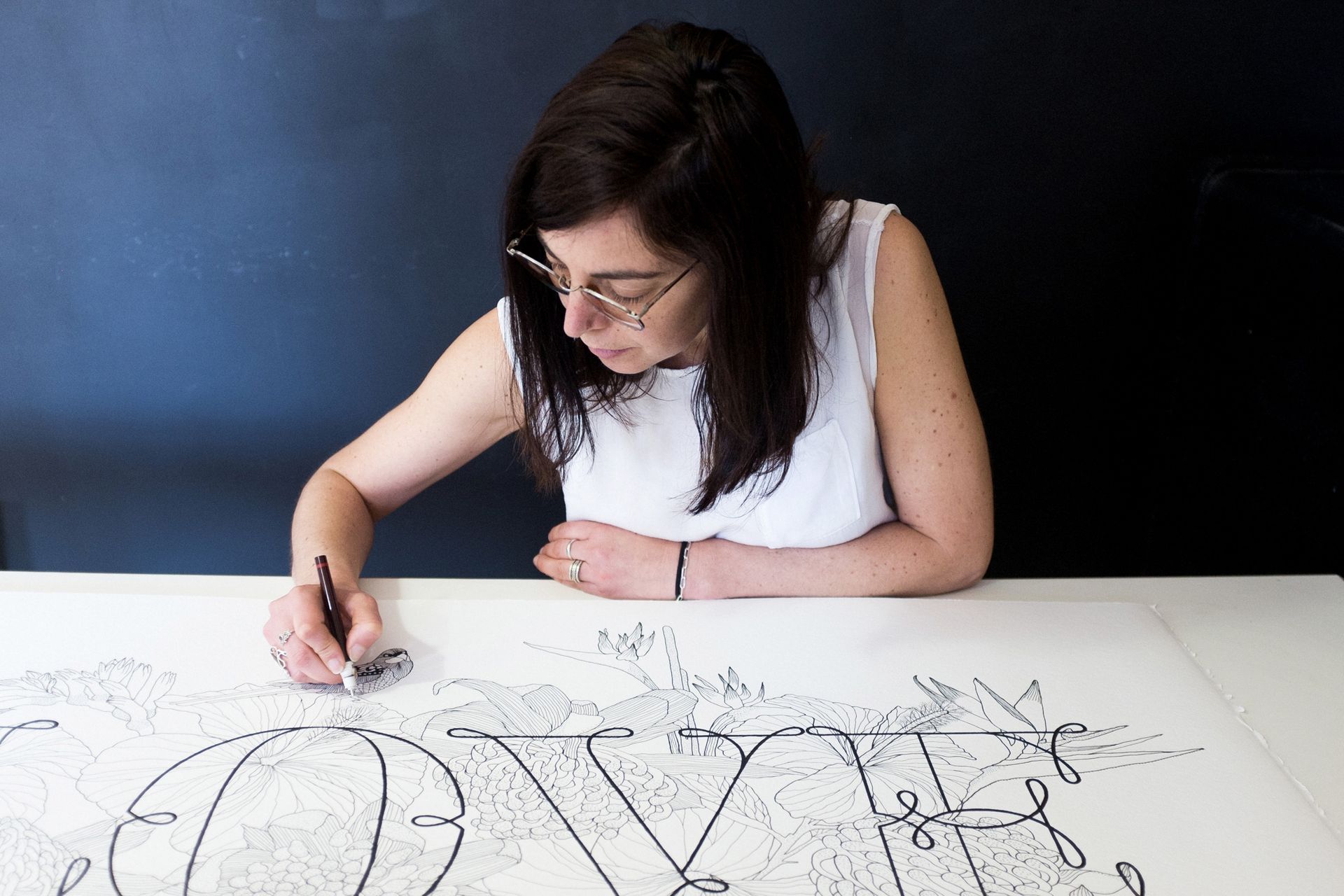 A person with dark hair and glasses meticulously draws ornate, black-ink script on a white surface against a dark background.