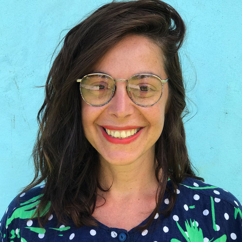 Smiling Maria Montes with glasses and dark hair against a turquoise wall, wearing a navy polka dot shirt