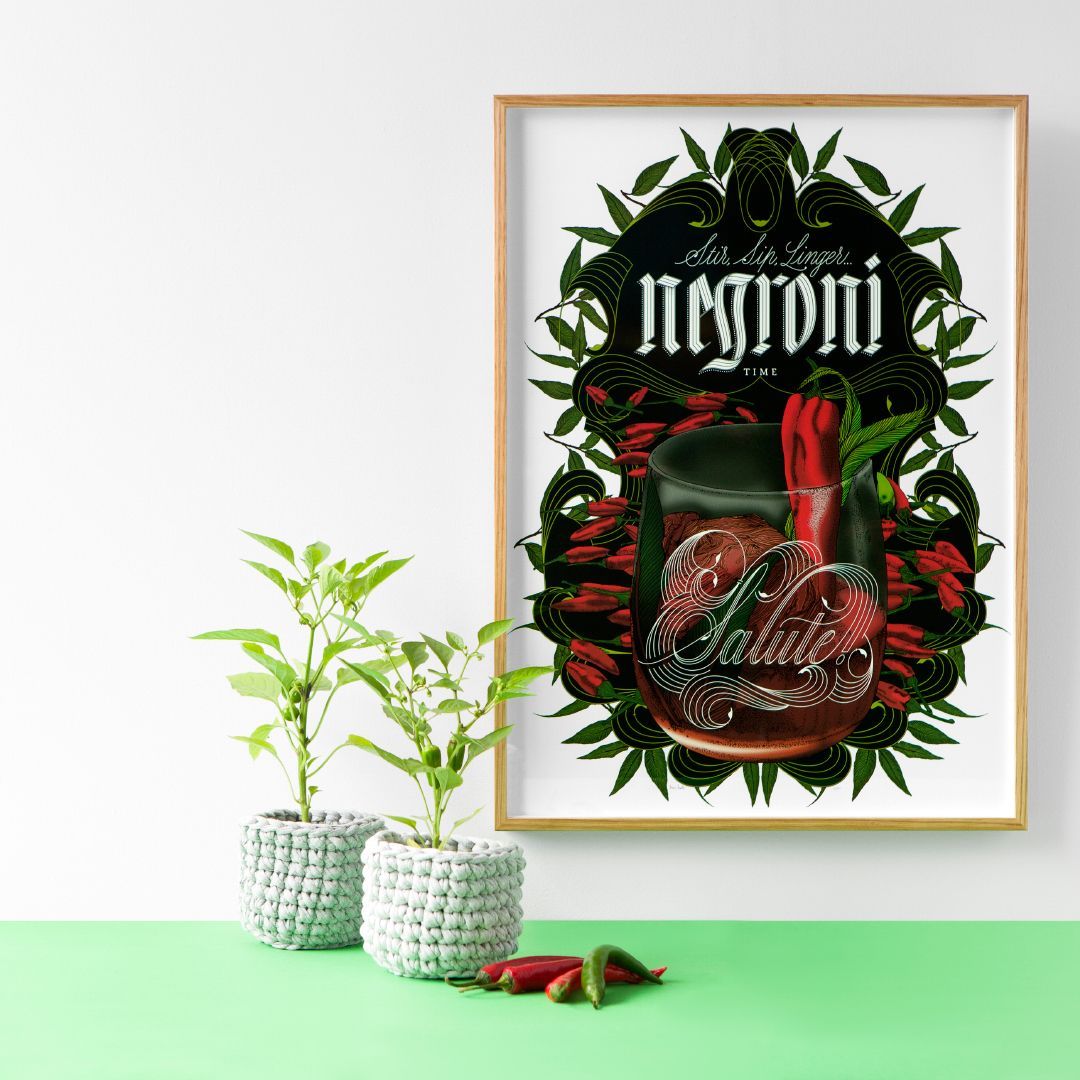 Framed Negroni cocktail poster beside two small potted plants on a mint-green surface