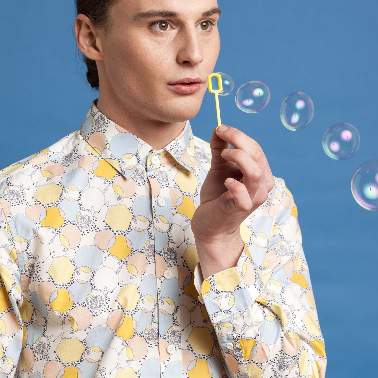 A person in a patterned yellow and blue shirt blows bubbles from a small yellow wand against a blue background.