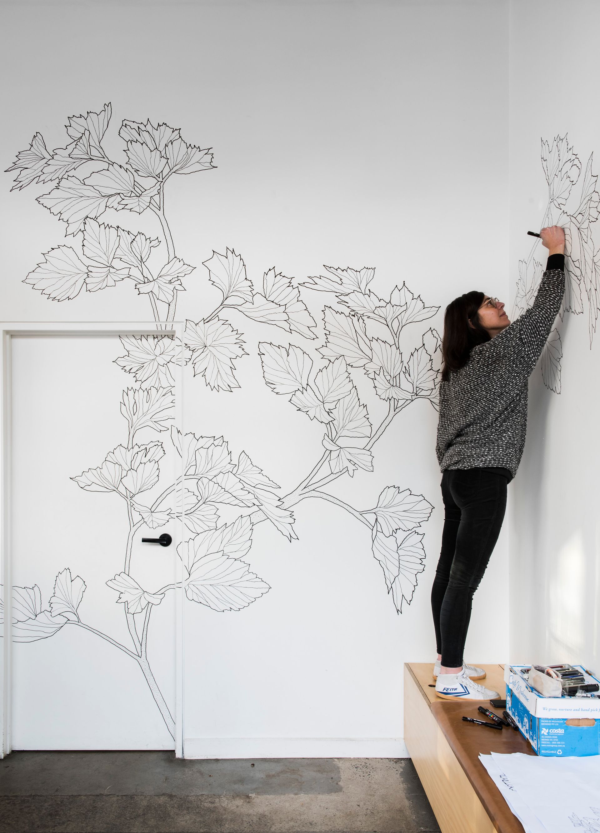 Person painting a black floral mural on a white wall beside a doorway.