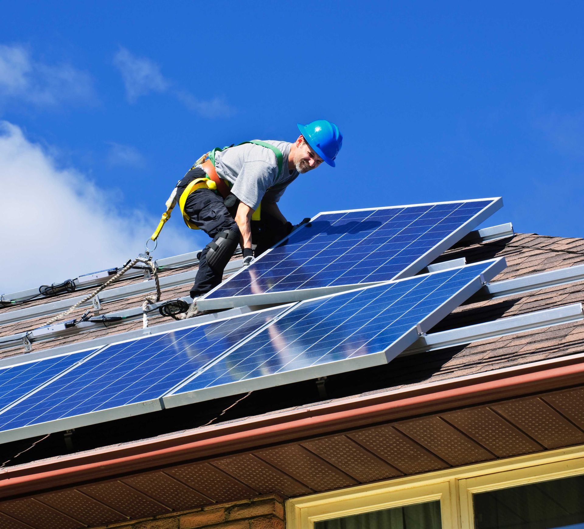 Un ouvrier, équipé d'un harnais de sécurité, installe des panneaux solaires sur un toit, sur fond de ciel bleu.
