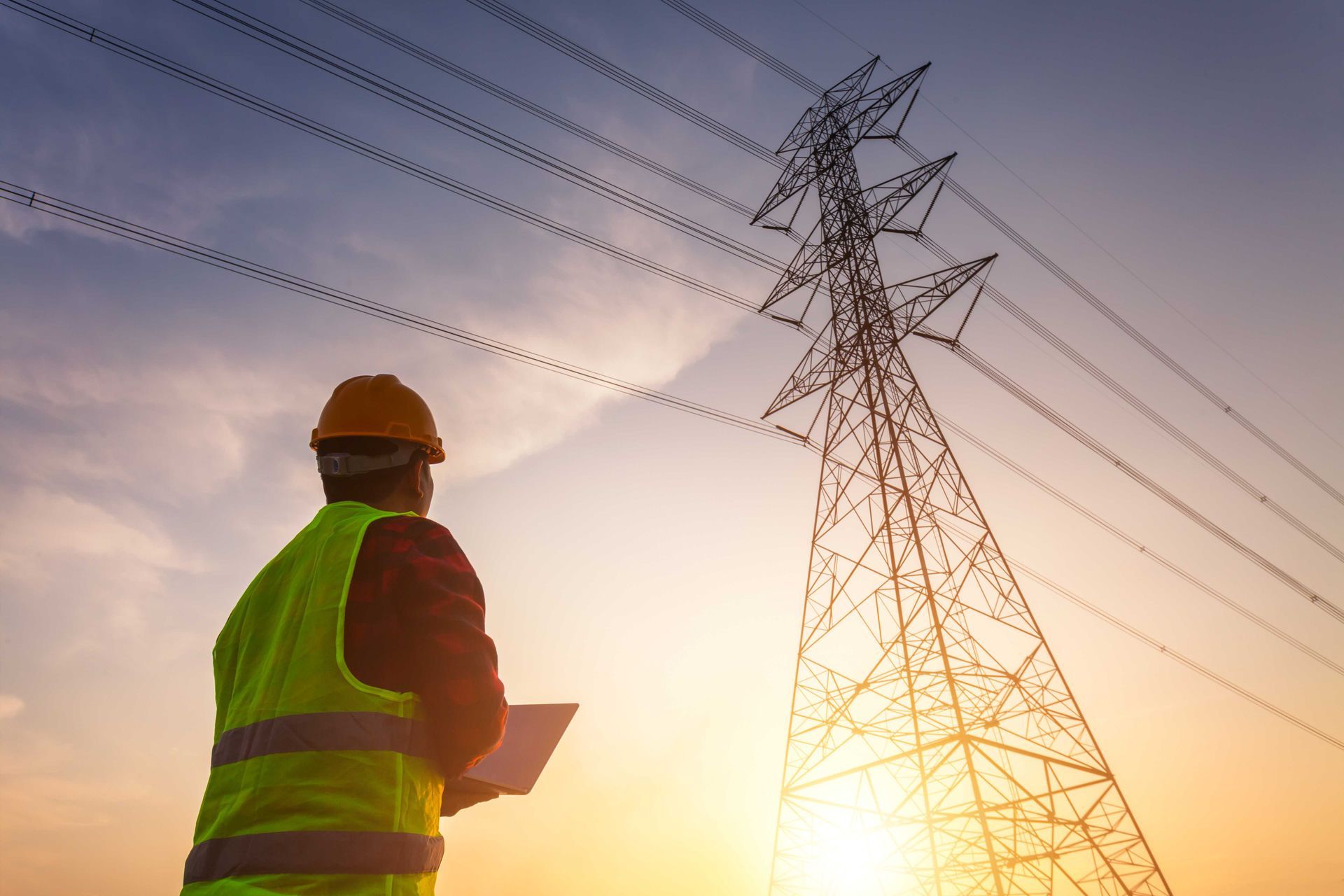 Un ingénieur, vêtu d'un gilet de sécurité et d'un casque de chantier, inspecte des lignes électriques près d'un pylône de transmission au coucher du soleil.