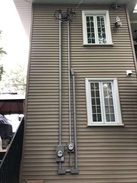 Extérieur de maison beige avec compteurs électriques, conduits et deux fenêtres à cadre blanc.