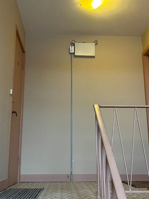 Couloir avec un boîtier blanc rectangulaire fixé en hauteur sur le mur, relié à un poteau métallique vertical.