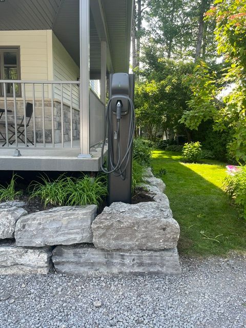 Borne de recharge pour véhicules électriques gris foncé, fixée sur un mur de pierre à côté d'une maison avec porche.