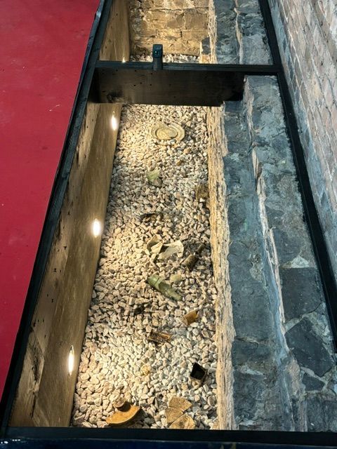 Plancher de verre révélant une tranchée éclairée et remplie de gravier entre des murs de briques et de béton ; bord rouge.