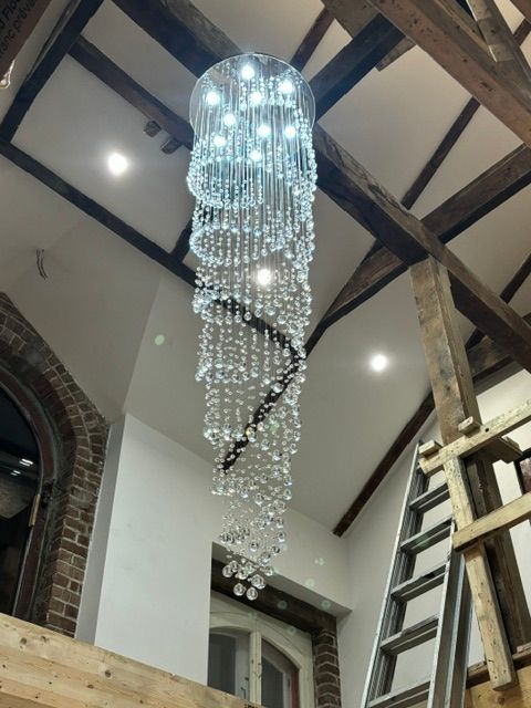 Lustre en cristal en spirale suspendu dans une pièce aux poutres apparentes, avec échelle et fenêtre.