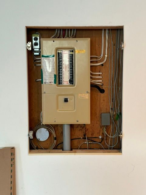 Tableau électrique encastré dans un mur, avec fils et conduits apparents. Panneau beige, cadre en bois clair, mur blanc.