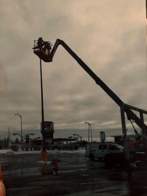 Une personne dans une nacelle élévatrice travaille sur un panneau de signalisation près d'une rue sous un ciel nuageux en hiver.