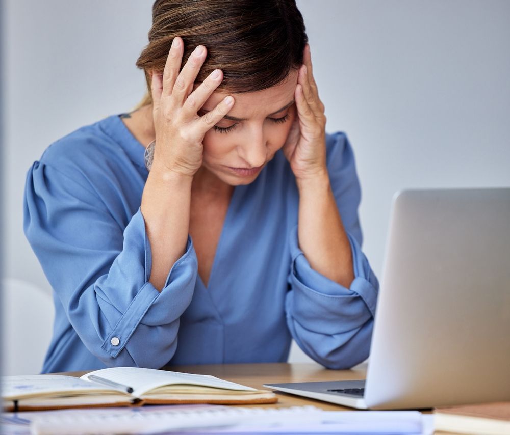 A woman is sitting at a desk with her hands on her head looking at a laptop.
