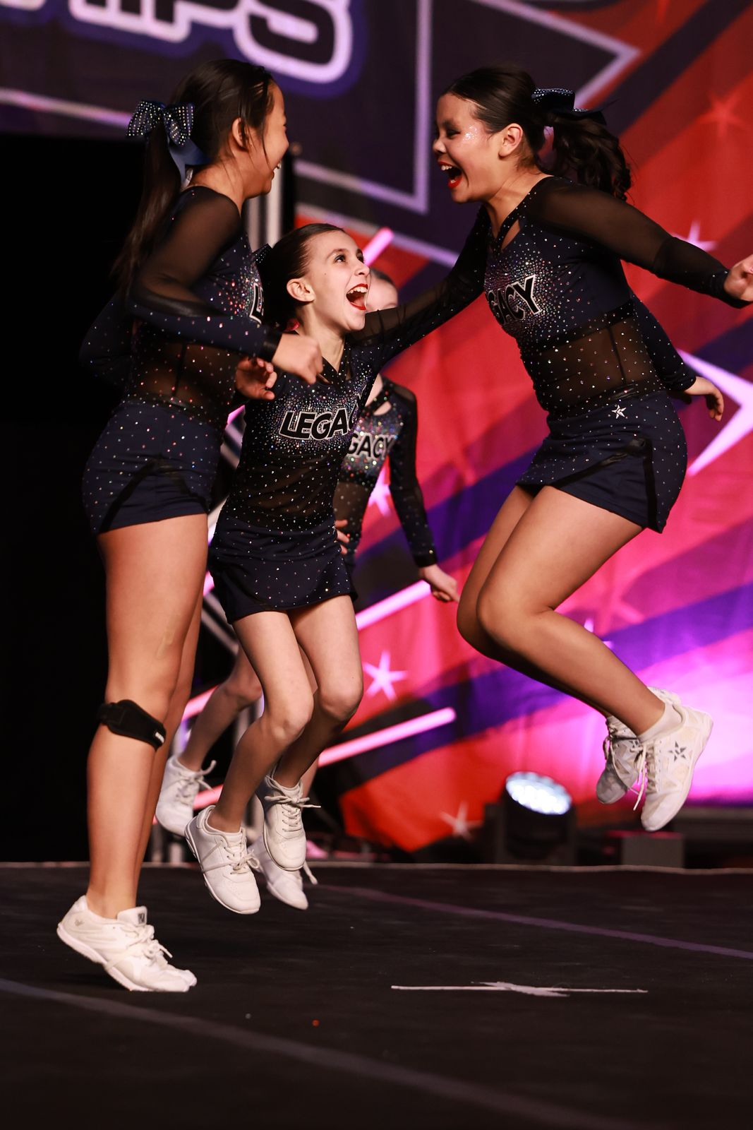 Three cheerleaders in black sparkly uniforms jumping on stage against a colorful backdrop