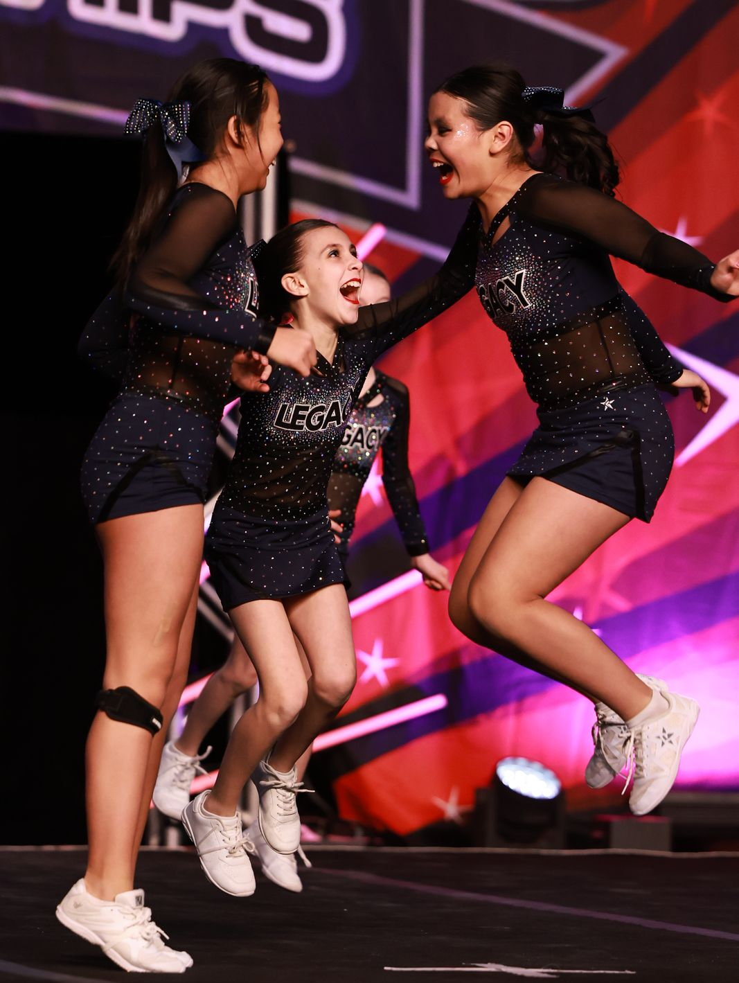 Three cheerleaders in black sparkly uniforms jump and smile on a stage