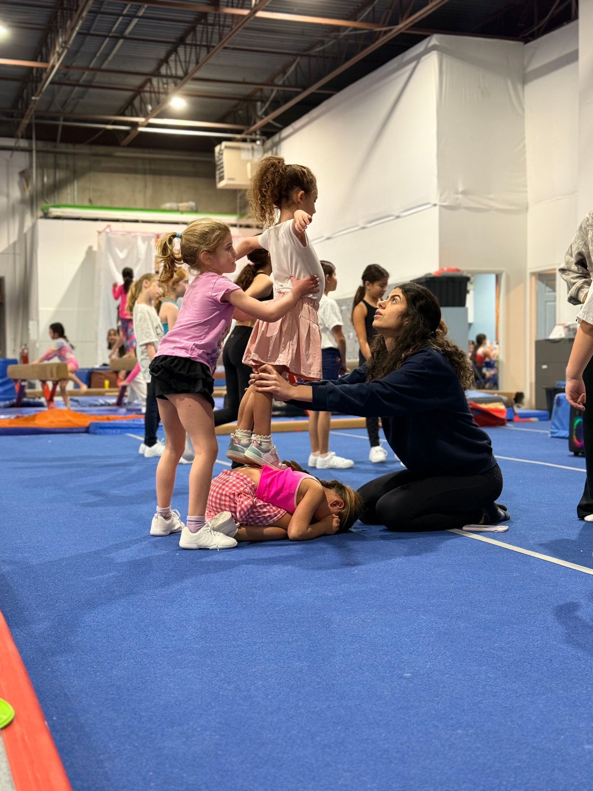 Children and adults in a gym, practicing tumbling on blue mats with a coach assisting nearby