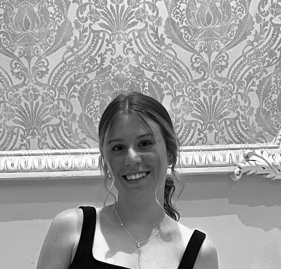 Smiling woman in a dark top, standing before ornate patterned wallpaper.