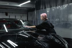 A woman is cleaning the windshield of a black car in a garage.