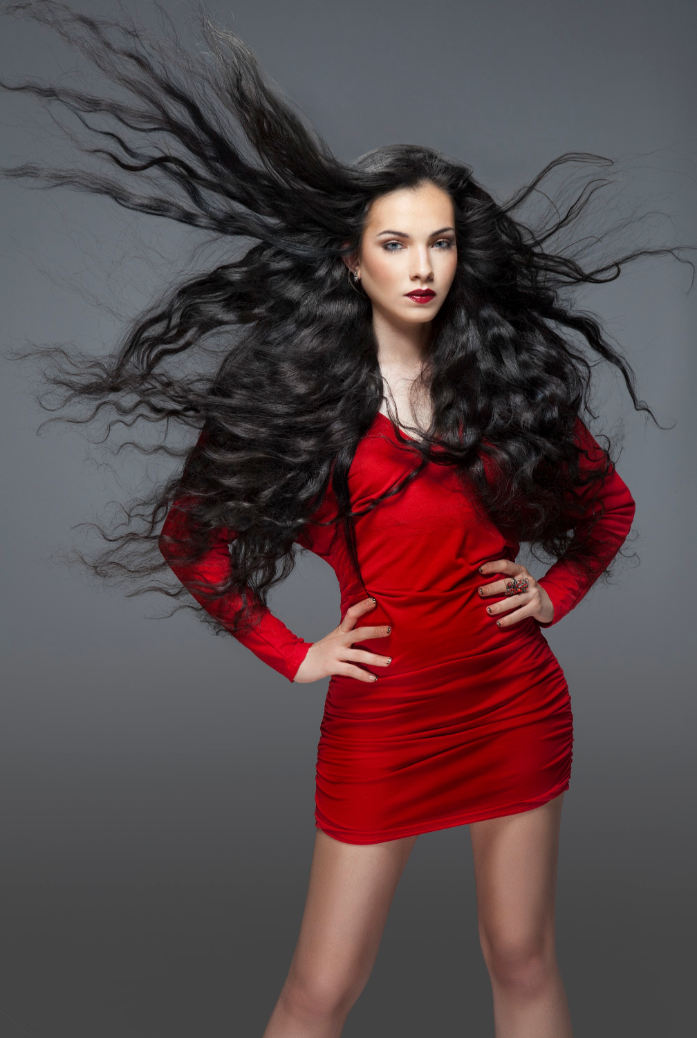 A woman in a red dress with long black hair is standing with her hands on her hips.