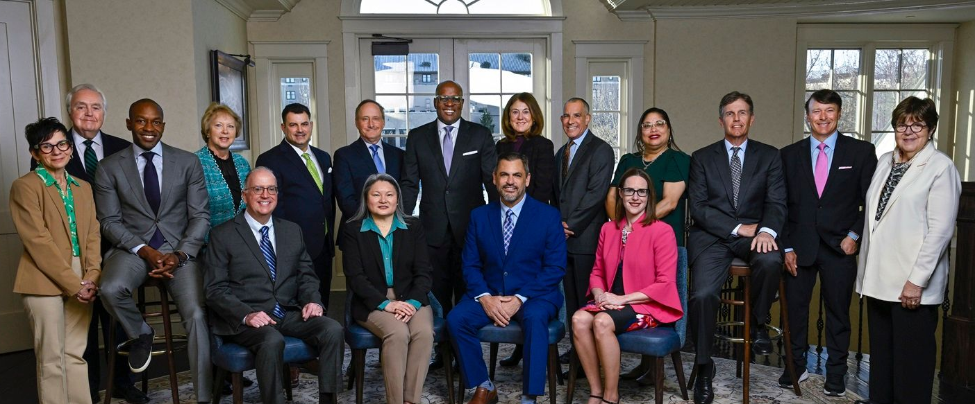 A group of 16 people in professional attire posing for a photo in an indoor setting with chairs and a large window.