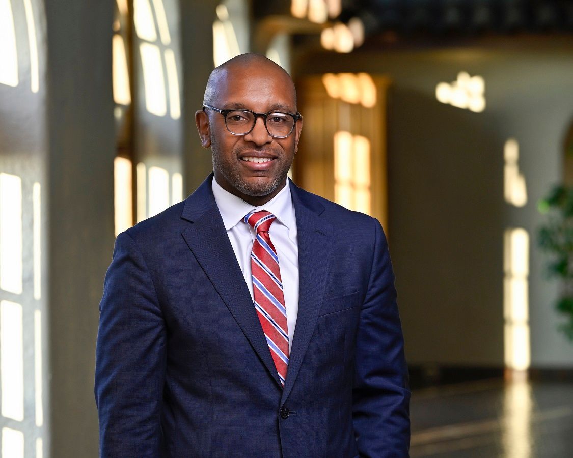 A man in a navy suit, white shirt, and red-striped tie smiling, posed in a blurred, sunlit hallway.