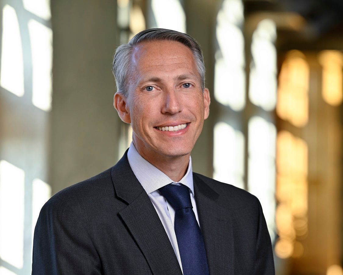 A smiling man in a dark suit and blue tie stands in front of a window with soft lighting.