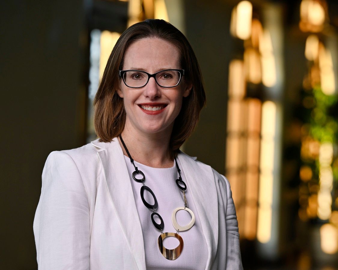 A person with glasses and a white blazer wears a large circular pendant necklace, smiling against a blurred indoor background.
