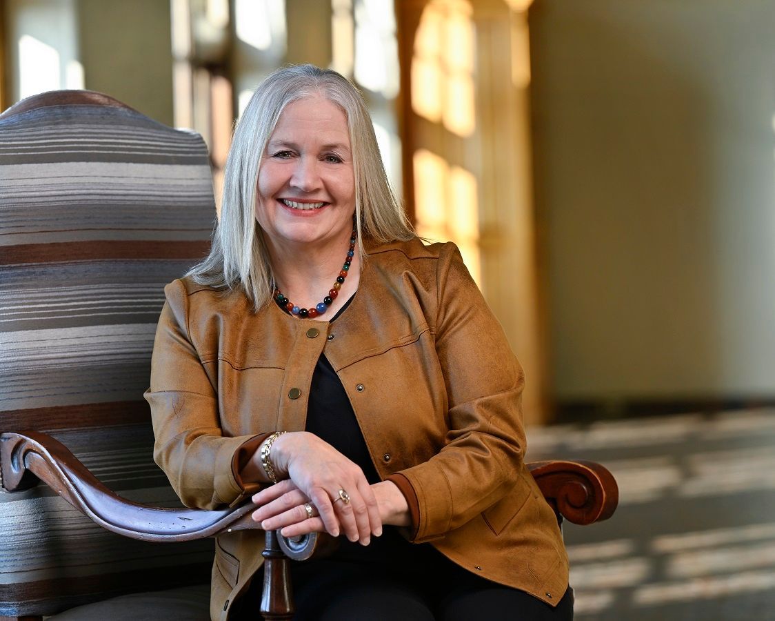A smiling person with shoulder-length gray hair wears a tan jacket and beaded necklace, sitting in a patterned chair.